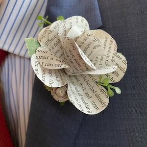 May include: A handmade paper flower boutonniere with text from a book. The flower is white with visible text and is accented with small green leaves and a pearl-like center. It is pinned to a dark blue suit.