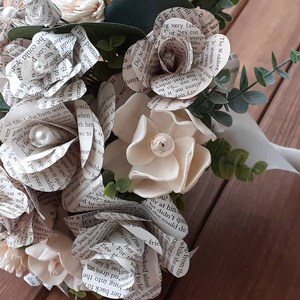 May include: A bouquet of white paper flowers made from book pages, with pearl accents, and greenery. The bouquet is tied with a white ribbon.