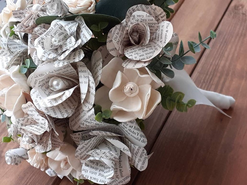 May include: A bouquet of white paper flowers made from book pages, with pearl accents, and greenery. The bouquet is tied with a white ribbon.