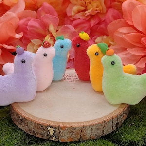 May include: A collection of colorful plush duck toys, each with a small fruit or vegetable on its head. The ducks are arranged on a wooden slice, with a backdrop of pink and orange flowers. The toys are in various colors.
