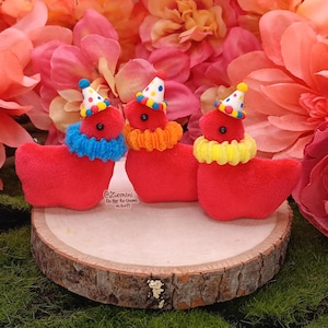 May include: Three red plush duck toys with party hats and colorful ruffled collars. The ducks are displayed on a wooden slice, surrounded by pink flowers and greenery. Each duck has a different colored collar: blue, orange, and yellow.