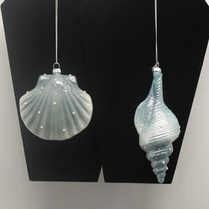 May include: Two blue and white glass Christmas ornaments shaped like seashells. The ornament on the left is a clam shell and the ornament on the right is a conch shell.