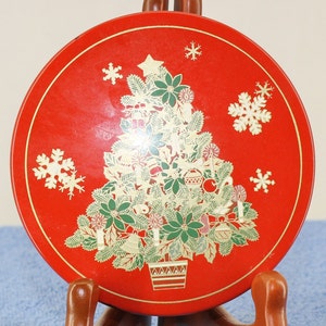 Puede incluir: Una lata redonda roja con un diseño de árbol de Navidad dorado. El árbol está decorado con adornos, campanas y velas. La lata tiene un diseño de copo de nieve dorado alrededor del borde.