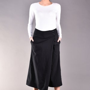 May include: A woman wearing a white long-sleeve shirt and a black wrap skirt. The skirt has a button closure and pockets. The woman is standing in front of a gray background.