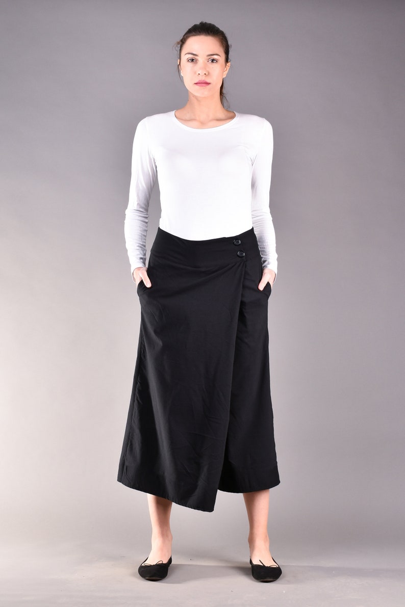 May include: A woman wearing a white long-sleeve shirt and a black wrap skirt. The skirt has a button closure and pockets. The woman is standing in front of a gray background.