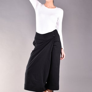 May include: A woman wearing a white long-sleeve shirt and black wrap-style culottes. The culottes have a button closure and a wide leg. The woman is also wearing black flats. The background is a solid gray.