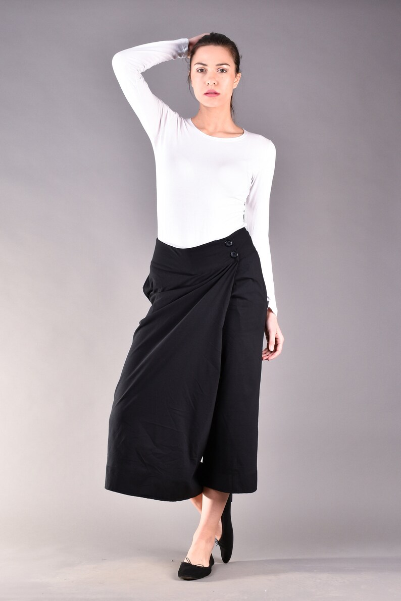 May include: A woman wearing a white long-sleeve shirt and black wrap-style culottes. The culottes have a button closure and a wide leg. The woman is also wearing black flats. The background is a solid gray.