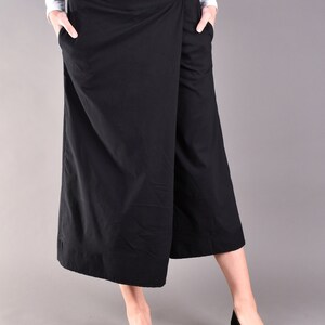 May include: Black wrap-style culottes with two buttons on the waistband and pockets. The model is wearing a white long-sleeve shirt and black flats. The culottes are knee-length and made of a solid black fabric.