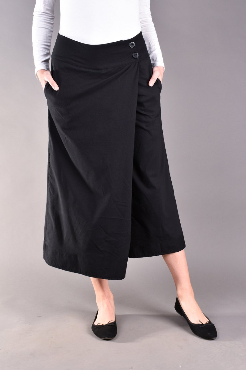 May include: Black wrap-style culottes with two buttons on the waistband and pockets. The model is wearing a white long-sleeve shirt and black flats. The culottes are knee-length and made of a solid black fabric.