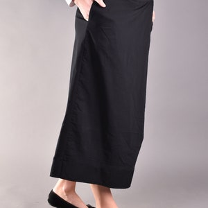May include: A long, black skirt with a high waist and two buttons on the side. The skirt has pockets and is paired with black flats. The model is wearing a white long-sleeve shirt.