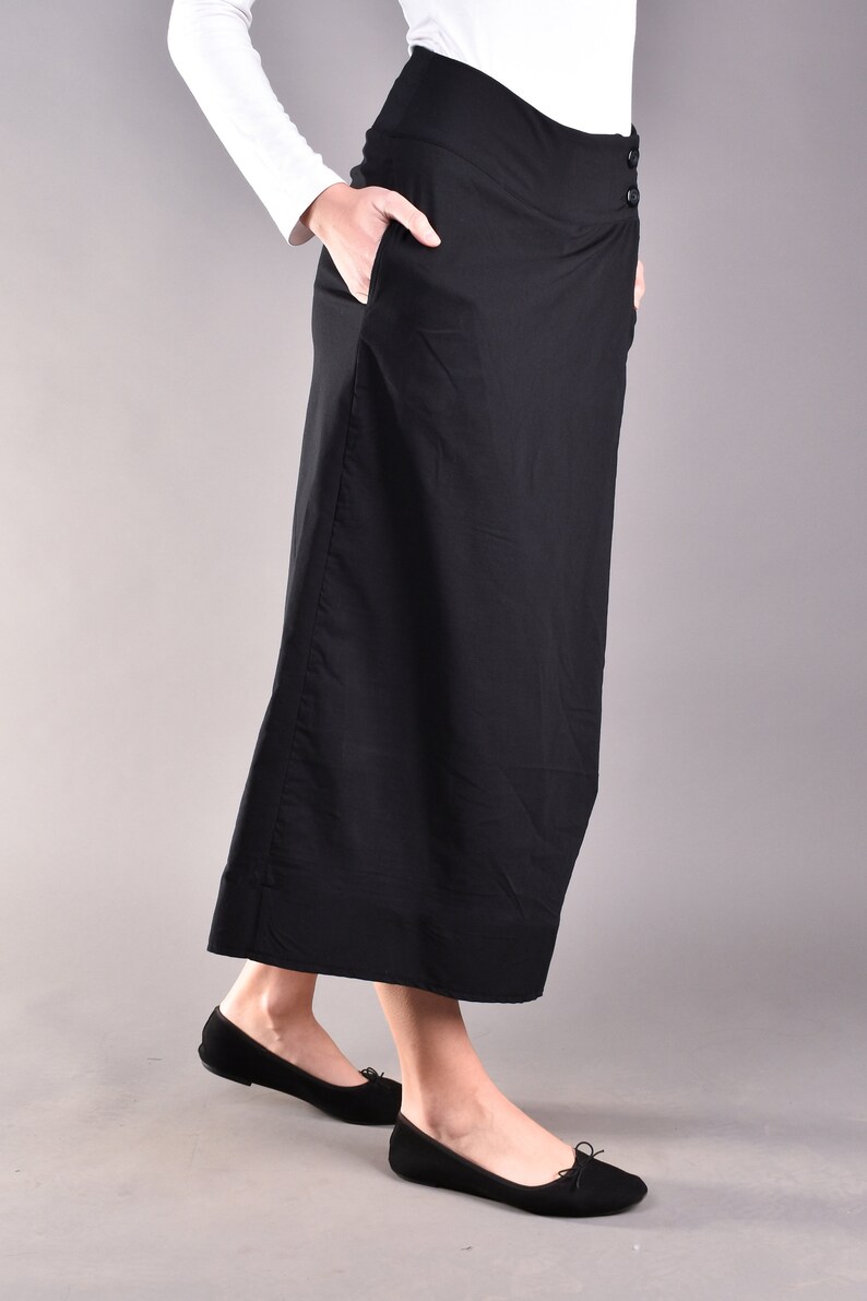May include: A long, black skirt with a high waist and two buttons on the side. The skirt has pockets and is paired with black flats. The model is wearing a white long-sleeve shirt.