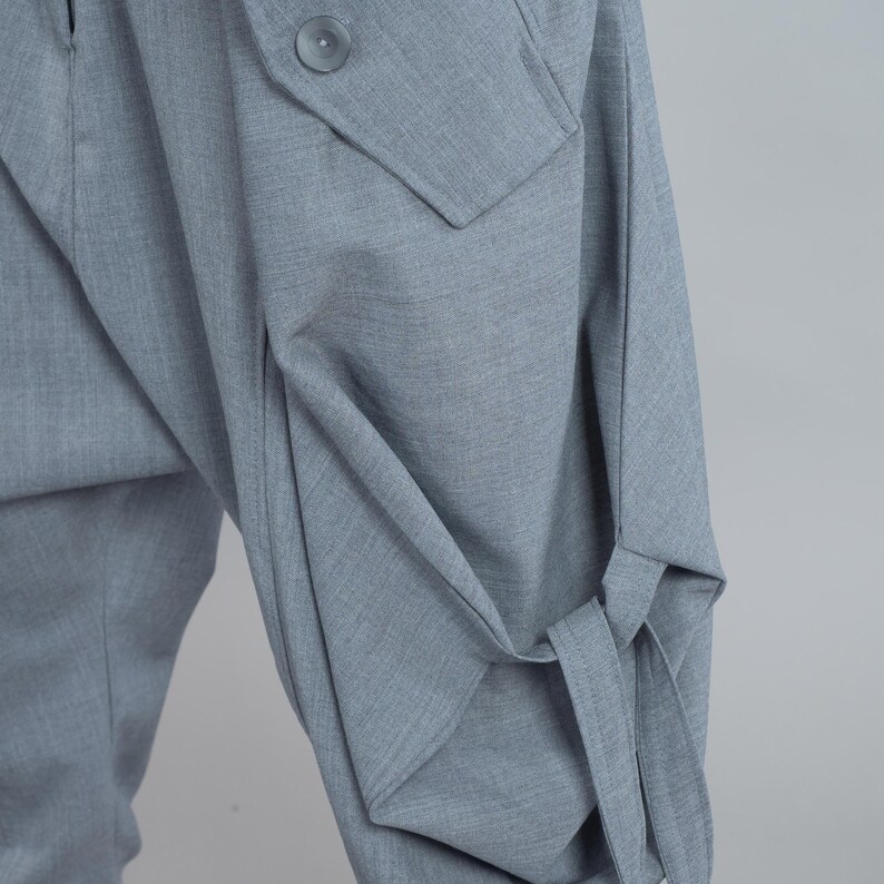 May include: Close-up of light grey tailored trousers with distinctive design features. Includes a buttoned pocket and fabric ties at the lower leg. The fabric has a subtle texture, suggesting a high-quality material.