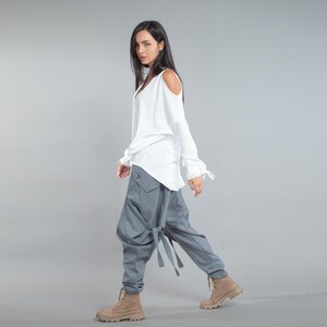 May include: A white top with cold shoulder cutouts and a V-neck is worn with grey, loose-fitting trousers. The trousers feature large pockets and strap details. Tan boots complete the outfit, creating a modern, casual style.