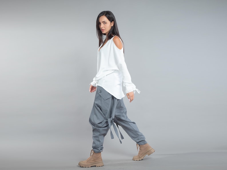 May include: A white top with cold shoulder cutouts and a V-neck is worn with grey, loose-fitting trousers. The trousers feature large pockets and strap details. Tan boots complete the outfit, creating a modern, casual style.