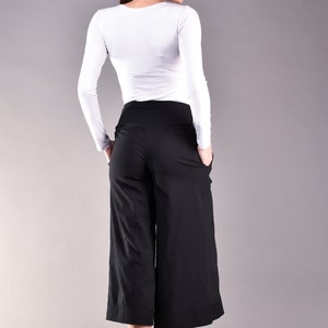 May include: A woman wearing a white long-sleeve shirt and black culottes. The culottes have a wide leg design and pockets. The woman is also wearing black flats. The background is a solid gray.