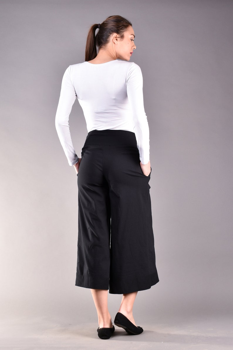 May include: A woman wearing a white long-sleeve shirt and black culottes. The culottes have a wide leg design and pockets. The woman is also wearing black flats. The background is a solid gray.