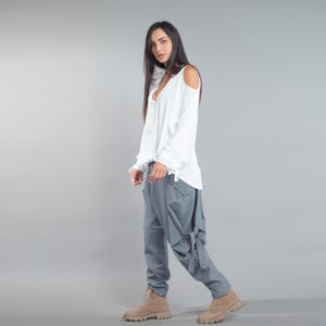 May include: A woman in a white long-sleeved top with shoulder cut-outs and grey trousers. The trousers feature a distinctive design with pockets and tie details. The outfit is completed with tan boots, creating a casual yet stylish appearance.