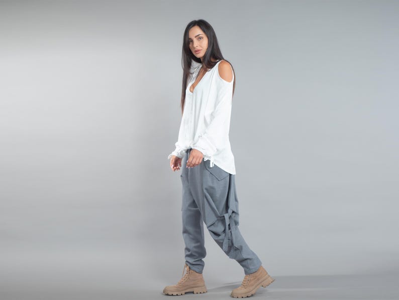May include: A woman in a white long-sleeved top with shoulder cut-outs and grey trousers. The trousers feature a distinctive design with pockets and tie details. The outfit is completed with tan boots, creating a casual yet stylish appearance.