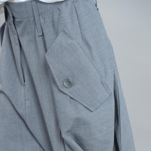 May include: Close-up of grey trousers with a large pocket on the side. The pocket has a button closure. The trousers are made of a textured fabric and have a pleated design.
