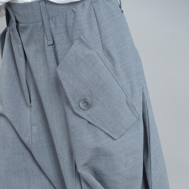 May include: Close-up of grey trousers with a large pocket on the side. The pocket has a button closure. The trousers are made of a textured fabric and have a pleated design.