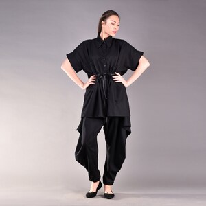 May include: A black, loose-fitting shirt with a collar and short sleeves. The shirt has a drawstring at the waist and a slightly asymmetrical hem. The model is wearing black pants with a loose, flowing fit.