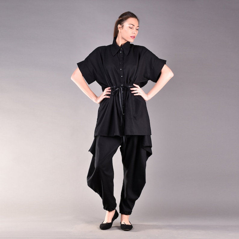 May include: A black, loose-fitting shirt with a collar and short sleeves. The shirt has a drawstring at the waist and a slightly asymmetrical hem. The model is wearing black pants with a loose, flowing fit.