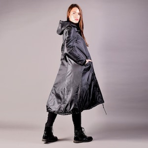 Peut inclure: Un long manteau noir à capuche avec un design fluide unique. Le manteau présente une taille cintrée et une jupe évasée, créant une silhouette distinctive. Le modèle porte des bottes et des leggings noirs. Le manteau a des poches et un ourlet à cordon.