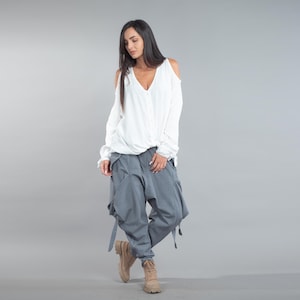 May include: A white top with a V-neck and cold shoulder detail, worn with grey cargo trousers with pockets and straps. The outfit is completed with tan boots. The trousers are a similar colour to the top.
