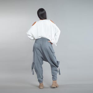 May include: A person seen from behind, wearing a white off-the-shoulder blouse and grey harem pants. The trousers have large pockets and straps at the knees. The person is wearing tan boots. The background is a solid grey.