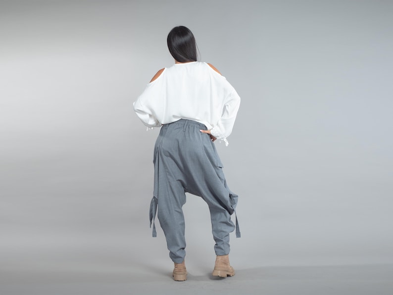 May include: A person seen from behind, wearing a white off-the-shoulder blouse and grey harem pants. The trousers have large pockets and straps at the knees. The person is wearing tan boots. The background is a solid grey.