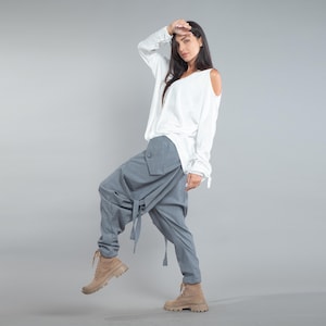 May include: A white long-sleeve top with a V-neck and cut-out shoulders paired with light blue harem pants. The pants have a unique wrap design and are tied at the waist. Tan boots complete the outfit. The clothing is displayed against a gray background.