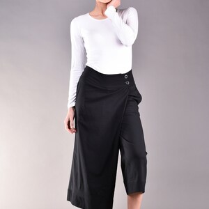 May include: Black culottes with a wrap-style front, featuring two button closures. The pants are paired with a white long-sleeve top and black flats. The outfit is set against a gray backdrop, showcasing a minimalist fashion aesthetic.