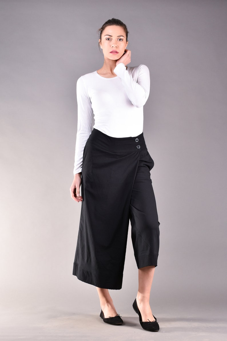 May include: Black culottes with a wrap-style front, featuring two button closures. The pants are paired with a white long-sleeve top and black flats. The outfit is set against a gray backdrop, showcasing a minimalist fashion aesthetic.