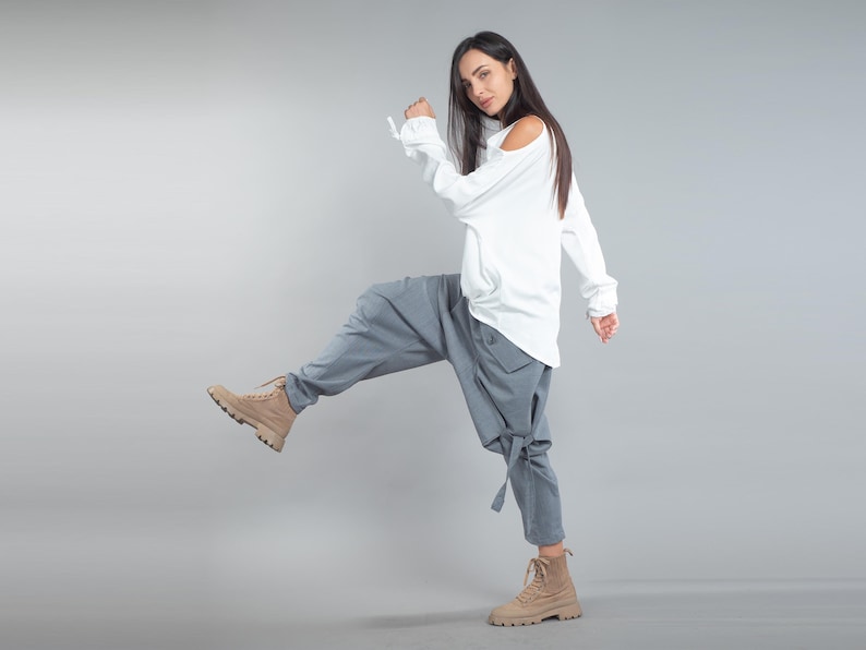 May include: A white long-sleeved top with a cold shoulder design, teamed with grey harem trousers and tan boots. The trousers have a unique, loose fit with tie details at the ankles. The outfit is set against a plain grey background.