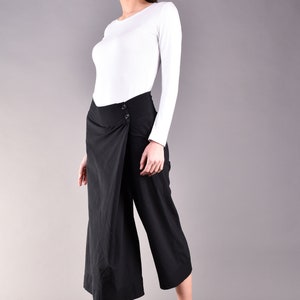 May include: A woman wearing a white long-sleeve shirt and black wrap-style culottes. The culottes have a button closure. She is also wearing black flats. The background is a solid gray.