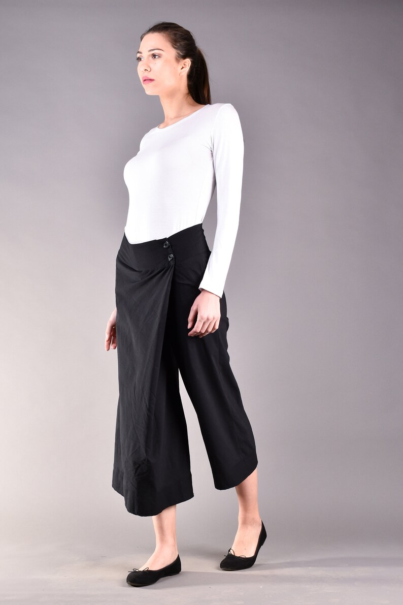 May include: A woman wearing a white long-sleeve shirt and black wrap-style culottes. The culottes have a button closure. She is also wearing black flats. The background is a solid gray.