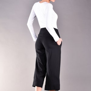 May include: A woman wearing a white long-sleeve shirt and black culottes. The culottes are knee-length and have pockets. The woman is also wearing black flats. The background is a solid gray.