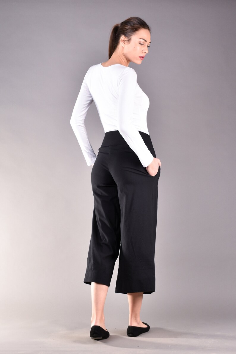 May include: A woman wearing a white long-sleeve shirt and black culottes. The culottes are knee-length and have pockets. The woman is also wearing black flats. The background is a solid gray.