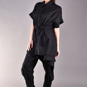 May include: A black shirt with a long, loose fit and a tie at the waist. The shirt has a collar and short sleeves. The model is wearing black pants and black shoes.