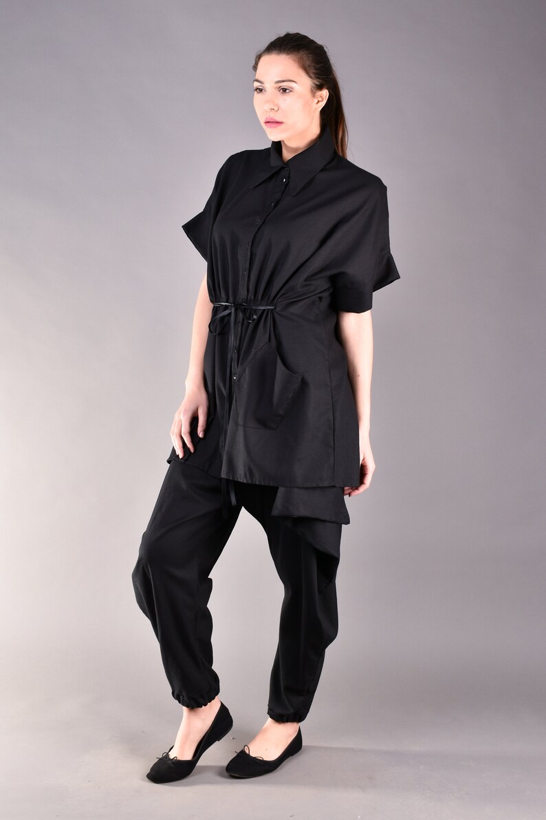 May include: A black shirt with a long, loose fit and a tie at the waist. The shirt has a collar and short sleeves. The model is wearing black pants and black shoes.