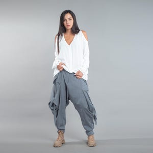 May include: A person is wearing a white, long-sleeved top with cold shoulders and a V-neck. They are also wearing loose-fitting, grey harem trousers and tan boots. The outfit is set against a grey backdrop.