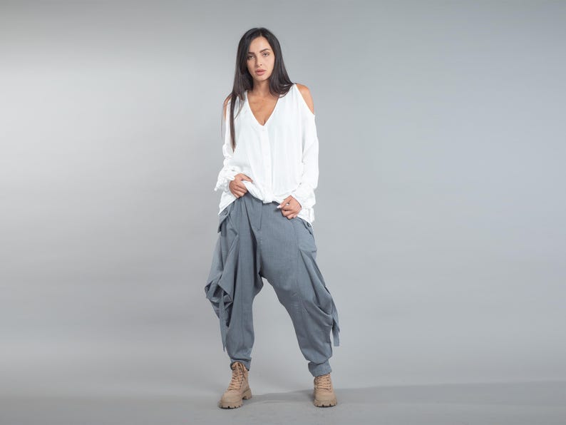 May include: A person is wearing a white, long-sleeved top with cold shoulders and a V-neck. They are also wearing loose-fitting, grey harem trousers and tan boots. The outfit is set against a grey backdrop.