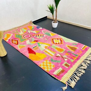 May include: A pink and multi-colored rug with a geometric pattern. The rug has a fringe border and is made of wool.