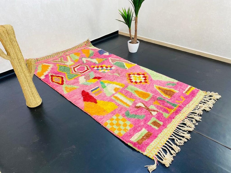May include: A pink and multi-colored rug with a geometric pattern. The rug has a fringe border and is made of wool.