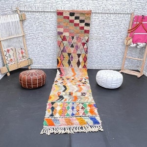 May include: A colorful Moroccan rug with geometric patterns in shades of pink, orange, yellow, blue, and green. The rug is laid out on a black floor with two poufs, one brown and one white.