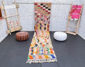 May include: A colorful Moroccan rug with geometric patterns in shades of pink, orange, yellow, blue, and green. The rug is laid out on a black floor with two poufs, one brown and one white.