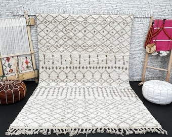 May include: A white and gray Moroccan rug with a geometric pattern. The rug has a fringe border and is laid out on a dark floor.