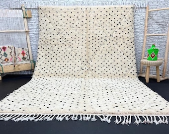 May include: A white and black Moroccan rug with a geometric pattern of small black dots. The rug has a fringe border and is laid out on a dark floor.