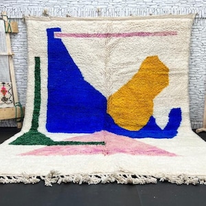 Puede incluir: Una alfombra blanca tejida a mano con formas geométricas en rosa, azul, verde y amarillo. La alfombra tiene un borde de flecos.
