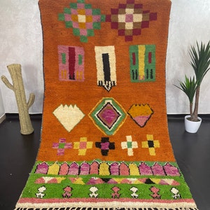 May include: A colourful Moroccan rug with geometric patterns in shades of orange, green, pink, and brown. The rug has a fringe border and is made of wool.
